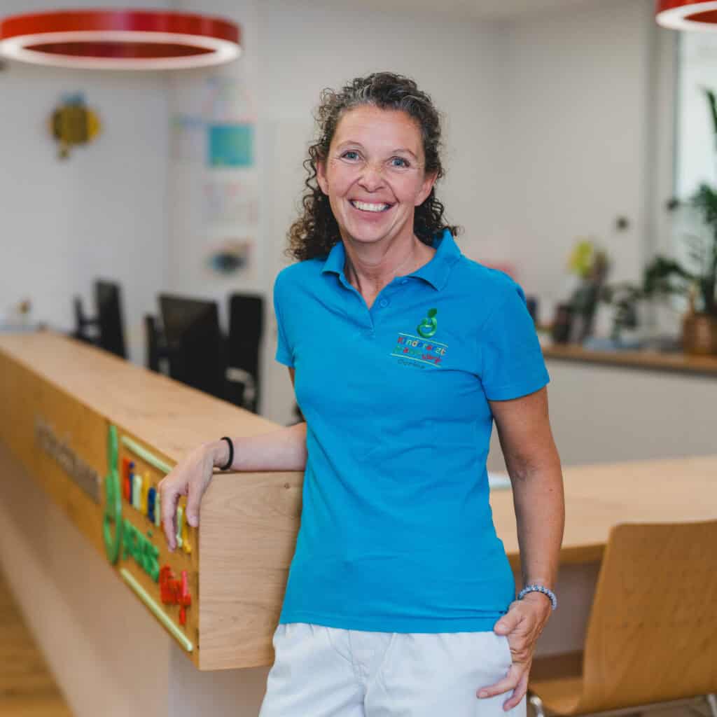 Carina lehnt am Tresen und lächelt freundlich. Sie trägt ein blaues T-Shirt.