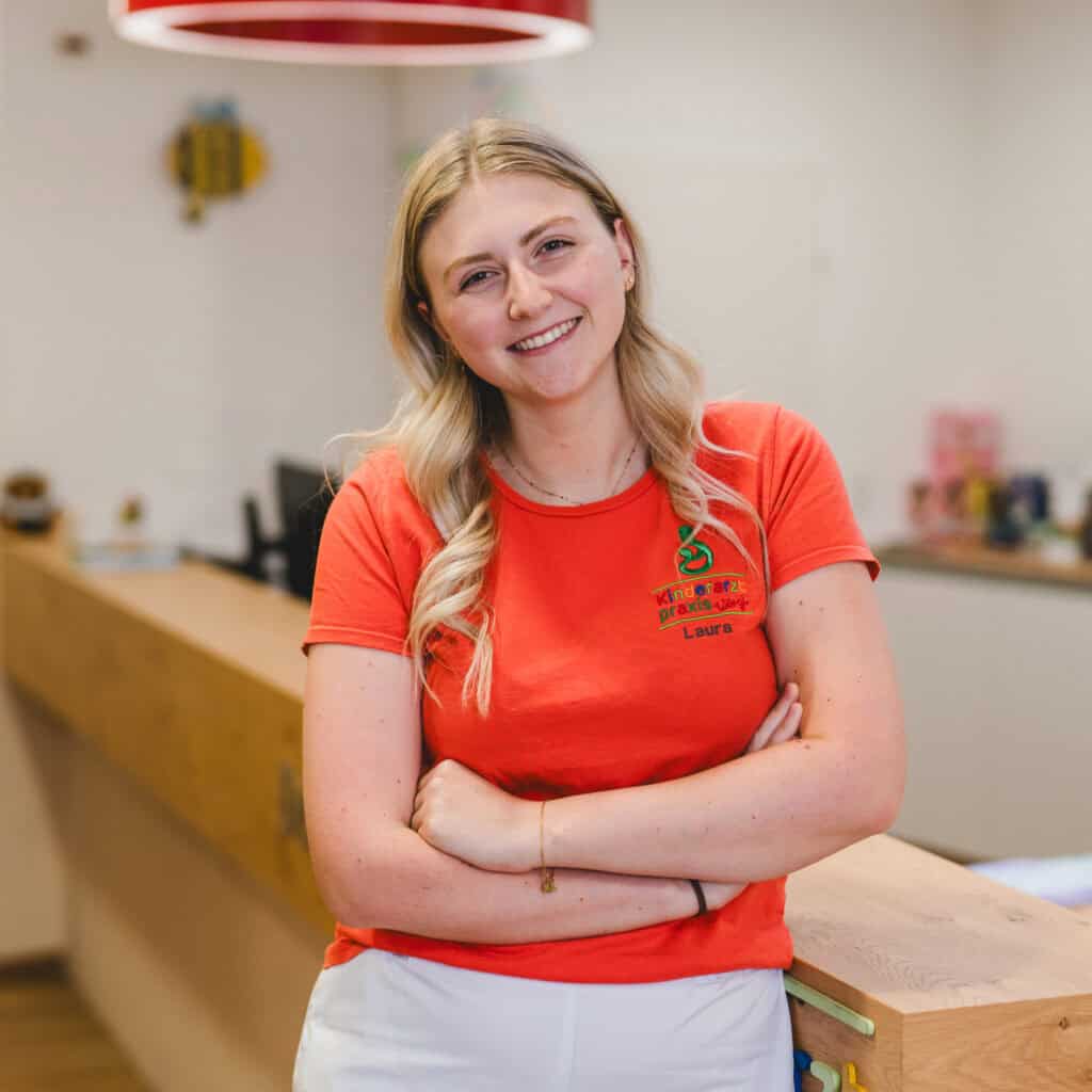 Laura lehnt am Tresen und hat ihre Arme verschränkt. Sie trägt ein apricot-farbenes Shirt.